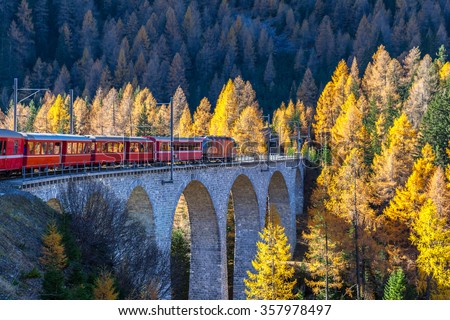 The sightseeing train bernina express of Rhaetian Railway running on the Viaduct with view of colorful trees on a sunny autumn day, Canton of Grisons, Switzerland