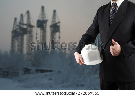 Close up of engineer hand holding white safety helmet for workers security, giving thumbs up success sign, standing in front of blurred urban background.