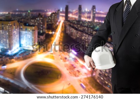 Close up of engineer hand holding white safety helmet for workers security standing in front of blurred urban background.