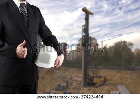 Close up of engineer hand holding white safety helmet for workers security, giving thumbs up success sign, standing in front of  blurred construction site in background