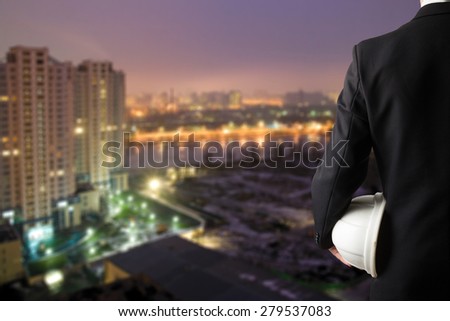 Close up of engineer hand holding white safety helmet for workers security standing in front of blurred urban background.