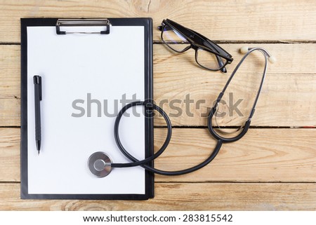Medical clipboard and stethoscope, glasses, black pen on wooden desk background. Top view. Workplace of a doctor.