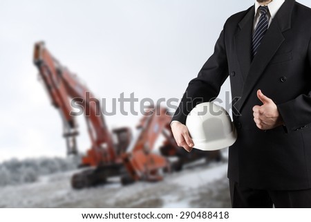 Close up of engineer hand holding white safety helmet for workers security standing in front of  blurred construction site with cranes in background