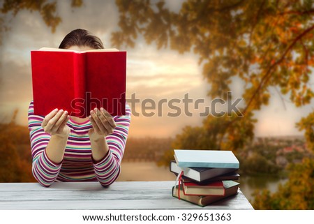 Young woman reading a book and covering her face ,sitting by wooden table with stack of colorful hardback books on blurred nature landscape backdrop.