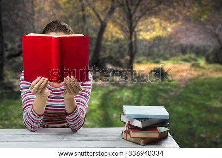 Young woman reading a book and covering her face ,sitting by wooden table with stack of colorful hardback books on blurred nature landscape backdrop. Copy space for text