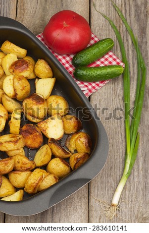 Fried potatoes in the pan on the aged wooden table with tomato, cucumbers and onion.