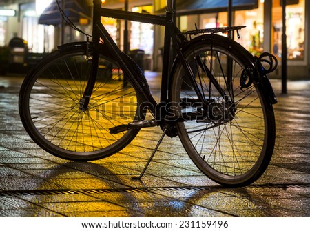 Wet bicycle parked on pavement with evening city lights in background