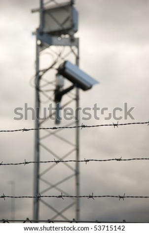 De-focused surveillance camera behind a fence with barbed wire. Focus on the fence.