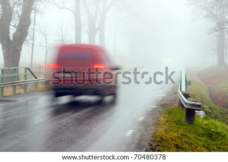 Red van driving on asphalt road in heavy fog