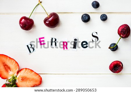 Berries Letters Cut out from Magazine with Ripe Cherries, Blueberries, Strawberries nearby on White Wooden Background