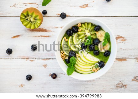 Healthy Green Smoothie in the Bowl from Banana and Spinach with Blueberries, Lime, Apple, Mint, Granola, Coconut and Kiwi on top, Top View