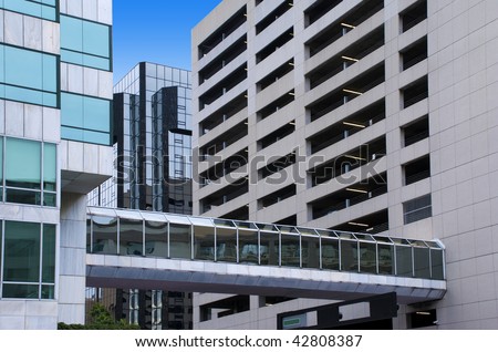 Elevated glass-covered city walkway between a tall office building and a tall parking garage.