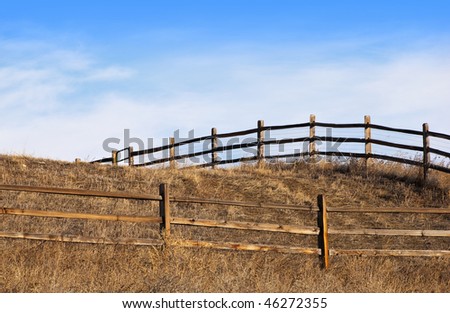 Wooden fence leads to the side and over a hilltop, with a gate partially visible, looking upward under a big, bright blue sky with wispy clouds