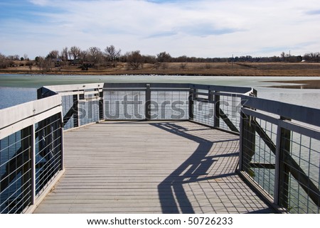 Small deck or pier on the edge of a small lake in flat country on the Colorado prairie, with view of some farm houses on the opposite side.
