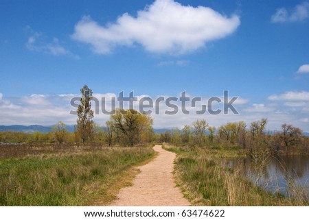 Open small dirt road or path leads straight ahead and forward, on a beautiful day through a wild area on the Colorado prairie, with water on one side.