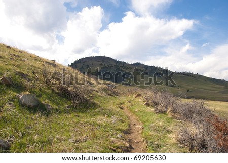 Path leads forward over a sunny hillside towards a shadowed hill beyond under dramatic clouds.