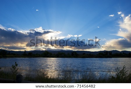 Sunset rays over golden clouds above a dark lake with ripples and grass silhouettes in front.