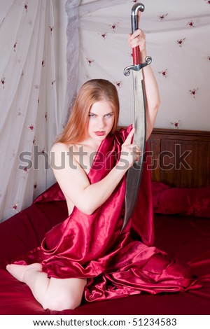 Gorgeous strawberry blond draped in red satin sheets holding a scimitar sitting on canopied bed