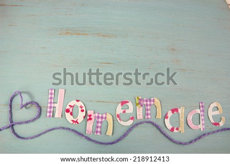 Fabric letters spell the word Homemade on a duck egg blue rustic distressed wood background. Underlined with a heart shape made from lilac yarn.background