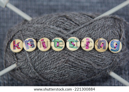 Grey ball of wool with the word knitting spelled in lettered buttons sewn to the wool. Knitting needles through wool and grey knitted background.
