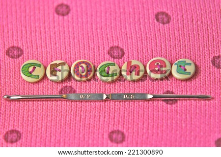 crochet spelled in lettered buttons on a pink wool background. Crochet hook underlines the word. Macro studio shot.