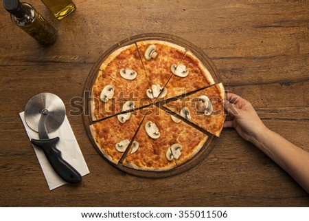 Top view of a sliced thin crusted mushroom Italian pizza on wooden table with pizza cutter and napkin. Hand taking slice of pizza.