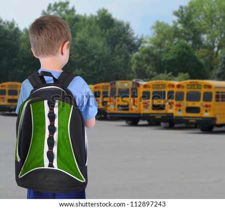 A young school boy is standing with a book bag and looking at school buses in the background.