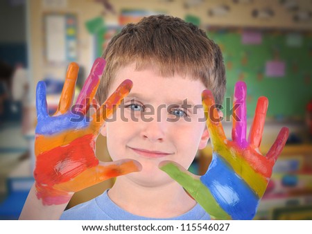 A small preschooler boy is holding up his hands with rainbow paint on his fingers in an art class.