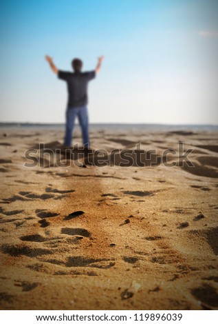 A man is standing on a beach with sand and has his hands held up in happiness, freedom or faith.
