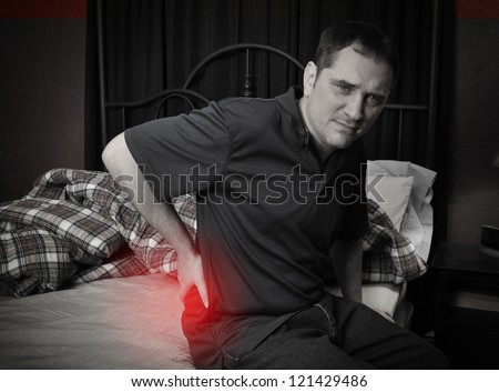 A man is sitting on his bed and rubbing his back in pain with a red glow to symbolize the ache.
