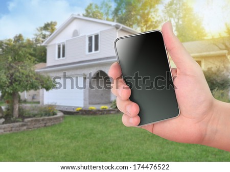 A hand is holding a smart phone in front of a house for a safety or control concept. The screen is blank to add your own message