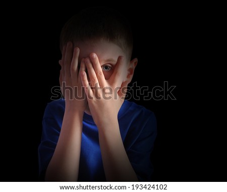 A child is hiding his eyes in the dark and looks scared or upset. The boy is isolated on a black background for a fear or sadness concept.