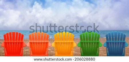 A rainbow of colors of wooden beach chairs are lined up along the water shore. There is copyspace in the clouds for a vacation message.