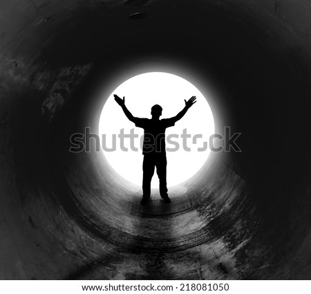 A person is at the end of a dark tunnel with a bright white light shining. The man has his hands in the air for a religious or freedom concept.