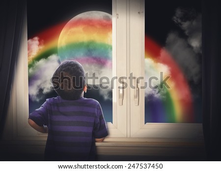 A young boy is looking out of the window in the night at a big moon with a rainbow for an imagination or creative concept.