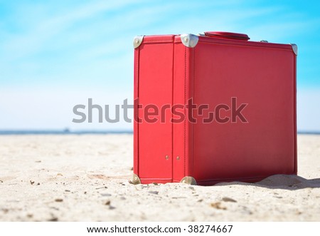 A red travel suitcase is alone on a beach with the lake or ocean in the background. Use this image to represent a voyage, getaway to a  tropical beach vacation in the sun.
