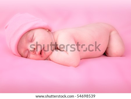A newborn baby child is laying down on a soft pink isolated background and sleeping with a hat on. The girl is laying belly down. Use the photo to represent life, parenting or childhood.
