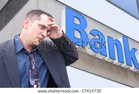 A business man is standing in front of a bank and looks stressed and worried. Can represent finance, the economy, or an investment theme.