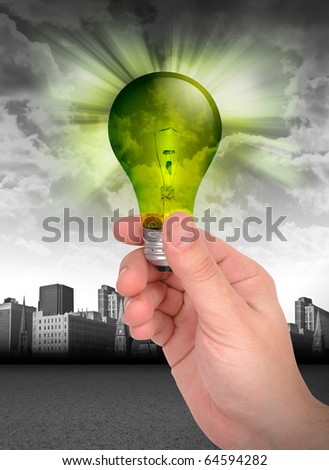 A hand is holding a green light bulb against a dark city. The lightbulb represents green energy and conservation. Can also be used as an idea or innovation concept.