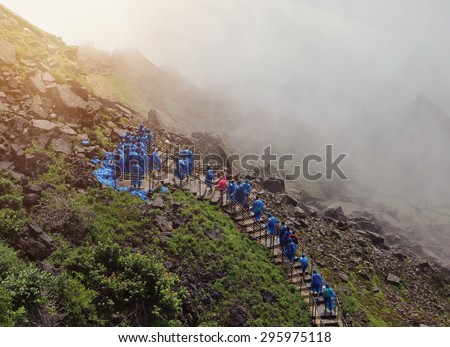 Tourists are walking up a stairway to Niagara Falls with wet water mist and blue raincoats on for a travel or nature concept.