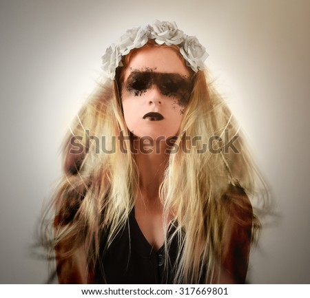 A scary dark woman has smokey black eyes and black lips with a flower headpiece and blonde hair blowing on a white background for a halloween or fear concept.