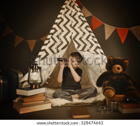 A boy is sitting in a tepee tent at night with  a light, books and teddy bear. He is looking up with a monocular for a exploration or camping concept.