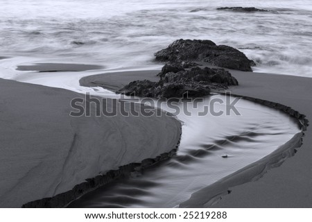a stream of water carves an inlet to the sea through the beach sand in Black and White