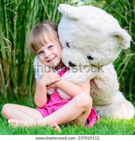 Little cute girl sitting in the grass with large teddy bear on her back
