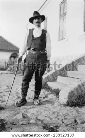 Serbian man wearing hat, vest, belted pants and pointed shoes with cane standing outside of building in Yugoslavia, ca 1880-1924.