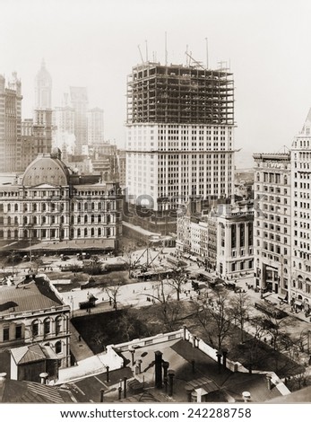 Woolworth Building, partially-constructed building amidst smaller buildings. The lower floors have received a white terra-cotta facing worked in lacy Gothic style. Photo made April 4th, 1912.