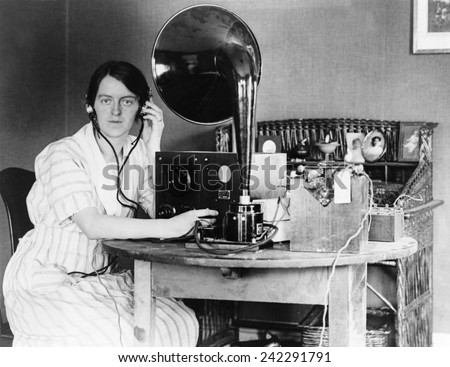 Woman listening to radio in her home receiving a personal radio message telling her when her husband will arrive home. 1922.