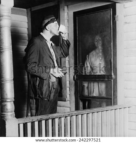 Homeless man in ragged clothes eating a meal standing as his benefactor keeps her distance behind her front door. 1919 photo by N.D. Hillis.