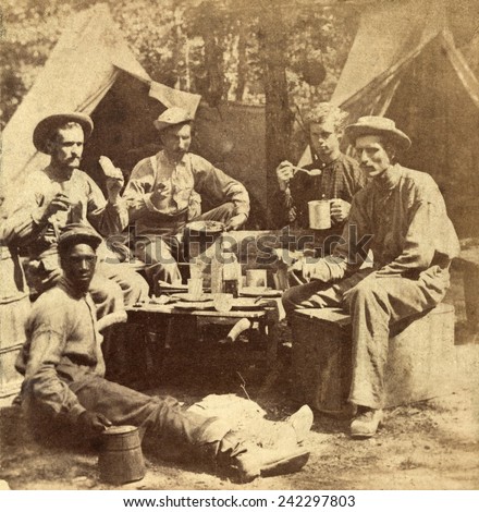 Relaxed scene of soldiers from the Army of the Potomac eat their meal in the company of an African American, probably a \'contraband,\' employed to cook and perform work for the soldiers. Ca. 1862-65.