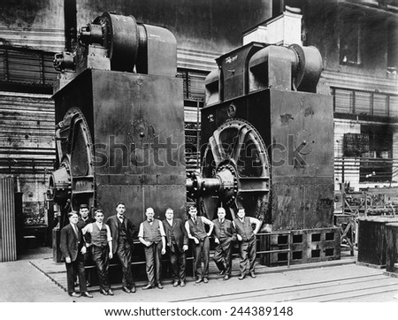 Nine men in suits are dwarfed by two massive dynamos, (electric generators) in a factory. Ca. 1920.